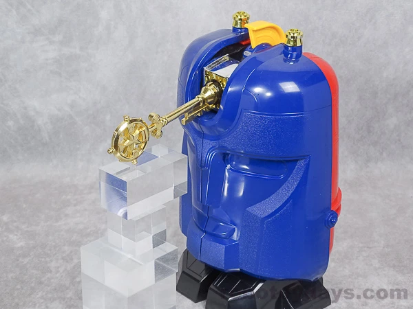 The gold key inserted into the forehead of DX Gunmazin's closed blue face, with clear display blocks supporting the accessory.